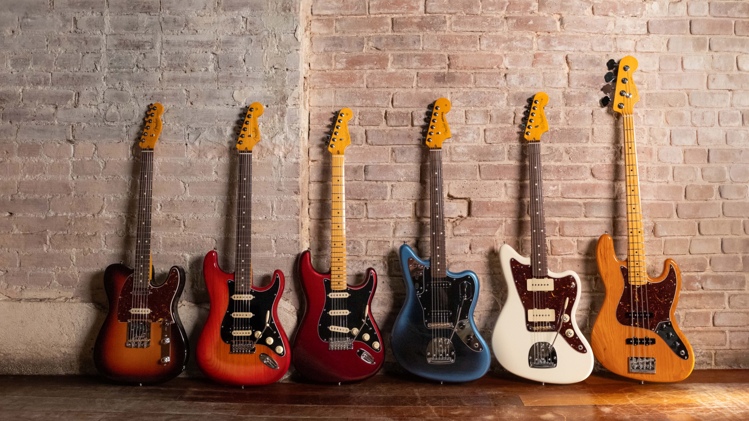 Six Fender American Professional guitars and basses lined up against a brick wall
