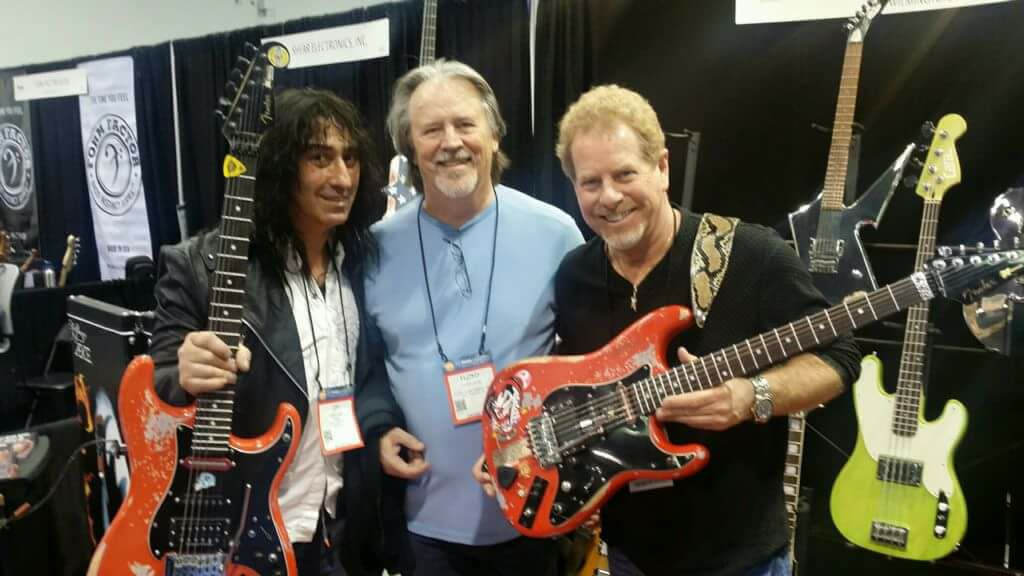 Three men smiling at a trade show booth; two of them hold electric guitars