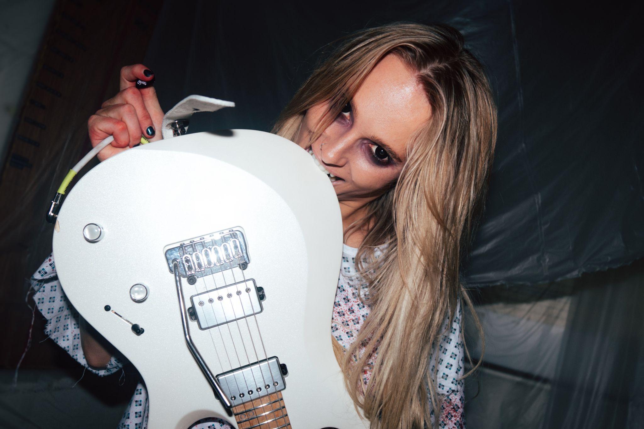 Sophie Lloyd holding a white electric guitar in a hospital gown in a press photo for her Avenged Sevenfold "Nightmare" guitar cover