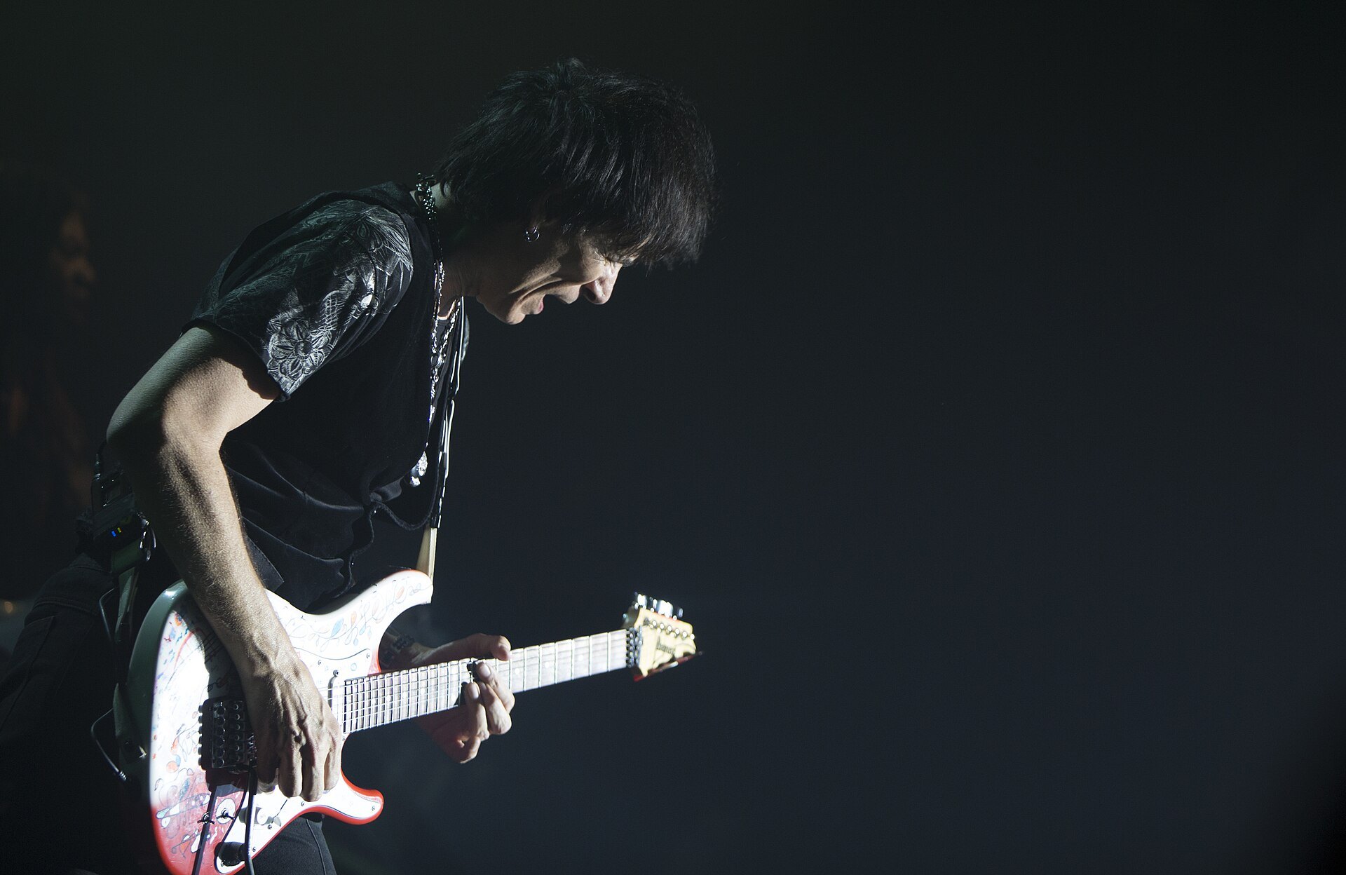 Steve Vai playing a white Ibanez guitar on a dark stage at Sala Barts in Barcelona