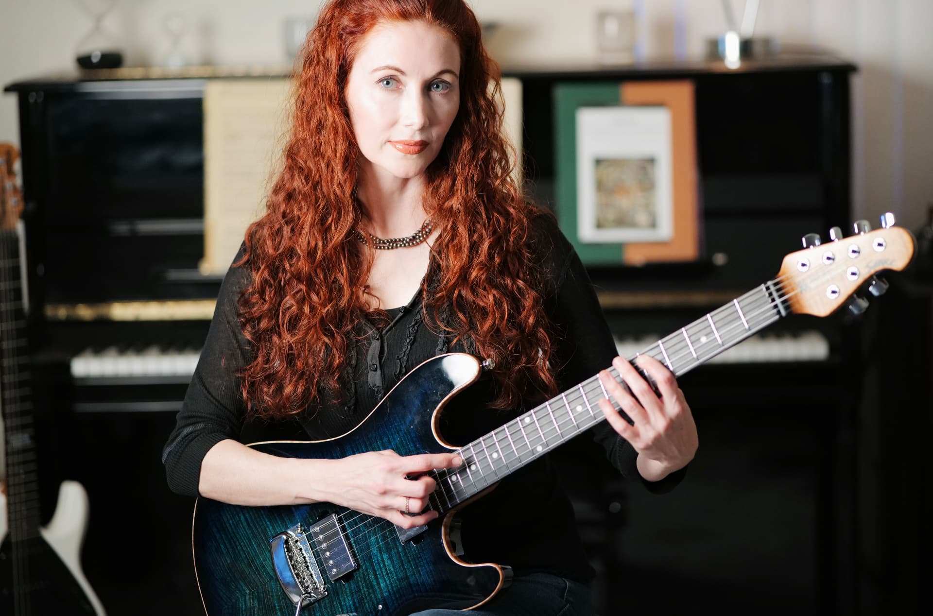 Gretchen Menn seated with a blue flame-top electric guitar in front of an upright piano and sheet music